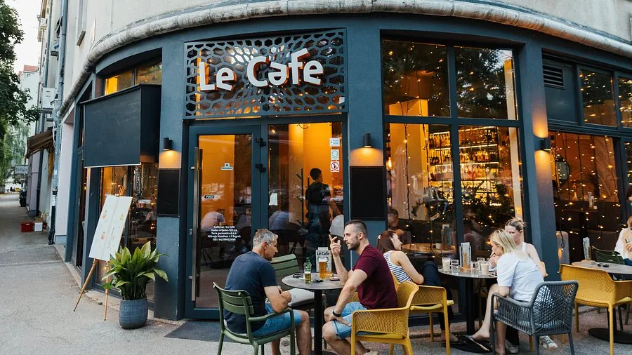 Teras sebuah cafe pada jam ramai menjelang malam, meja-meja luar terisi tamu dan bagian dalam terlihat menyala hangat